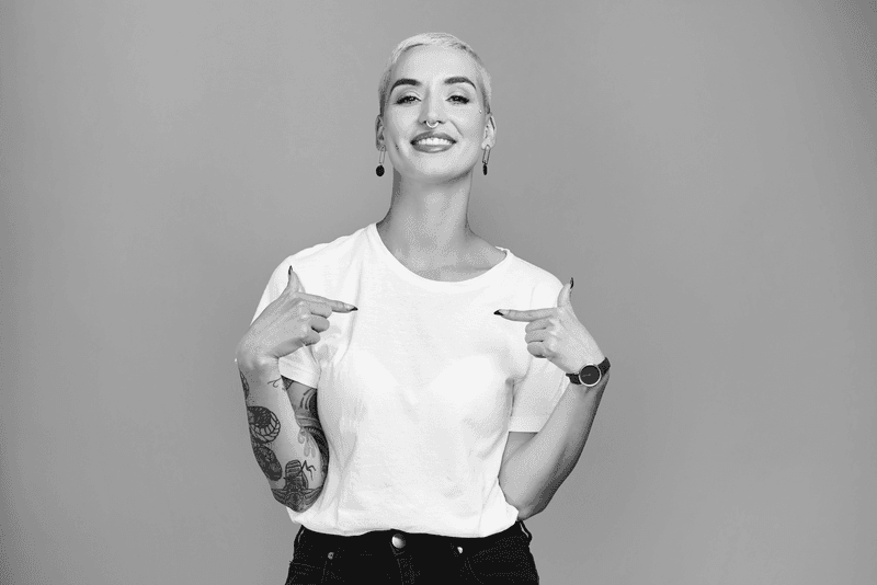 Smiling tattooed woman in a white t-shirt pointing to herself, representing our London coding bootcamps' empowering approach to learning.