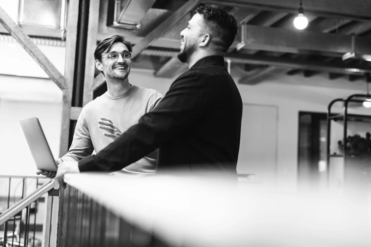 Two men leaning against a railing in an office space talk about talent acquisition.
