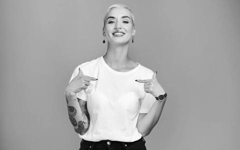 Smiling tattooed woman in a white t-shirt pointing to herself, representing our London coding bootcamps' empowering approach to learning.